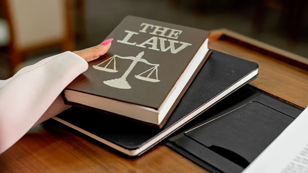 Close-up of a hand holding a book titled 'The Law' with legal documents and a notebook on a table.