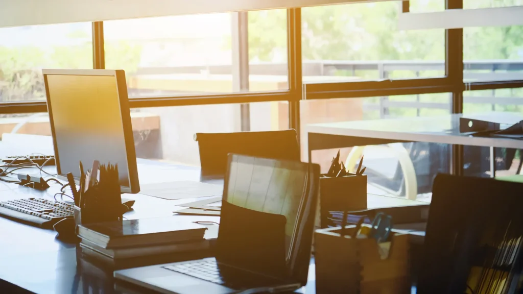 Modern office workspace with two laptops, pens, and office supplies on a desk, with sunlight streaming through large windows