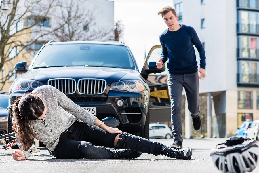 Man gets out of his car and runs to a woman laying and holding her leg in the street with her bicycle and helmet next to her