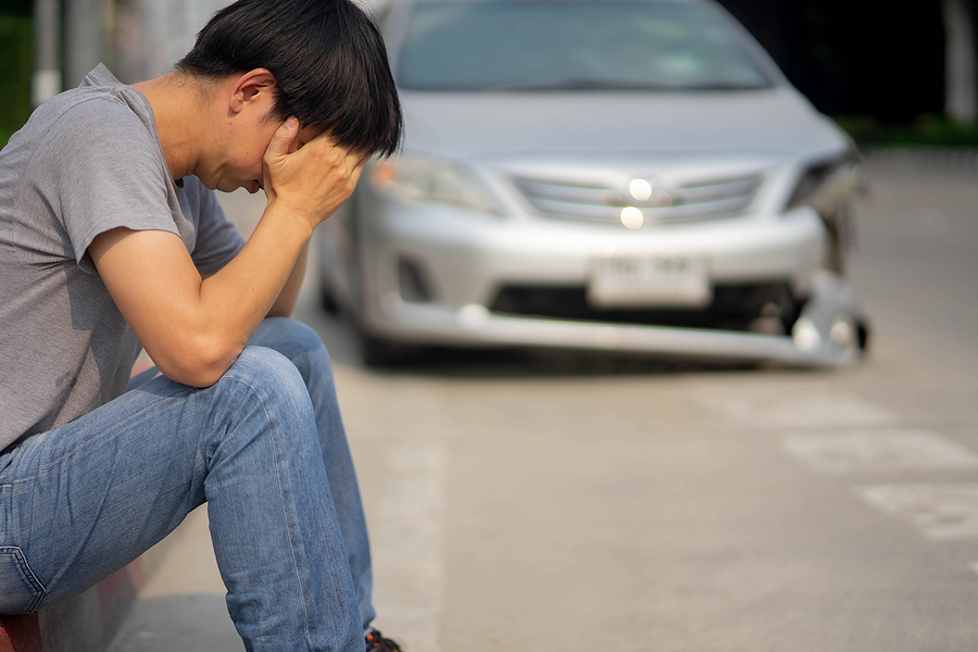 A man sits on the side of the road looking upset with his damaged car parked behind him