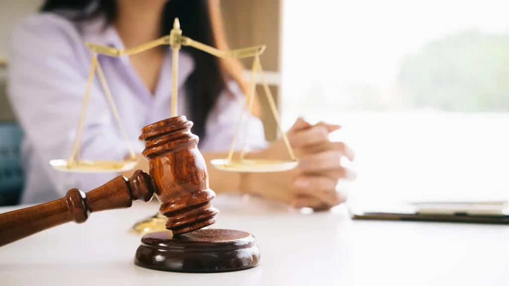 Close-up of a gavel and scales of justice on a table with a person’s hands folded in the background.